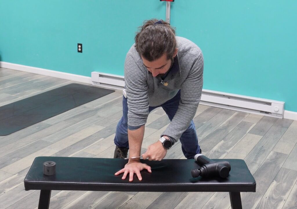 Dr. John with his hand on a bench using a muscle scraper in the crease of his wrist to break up scar tissue